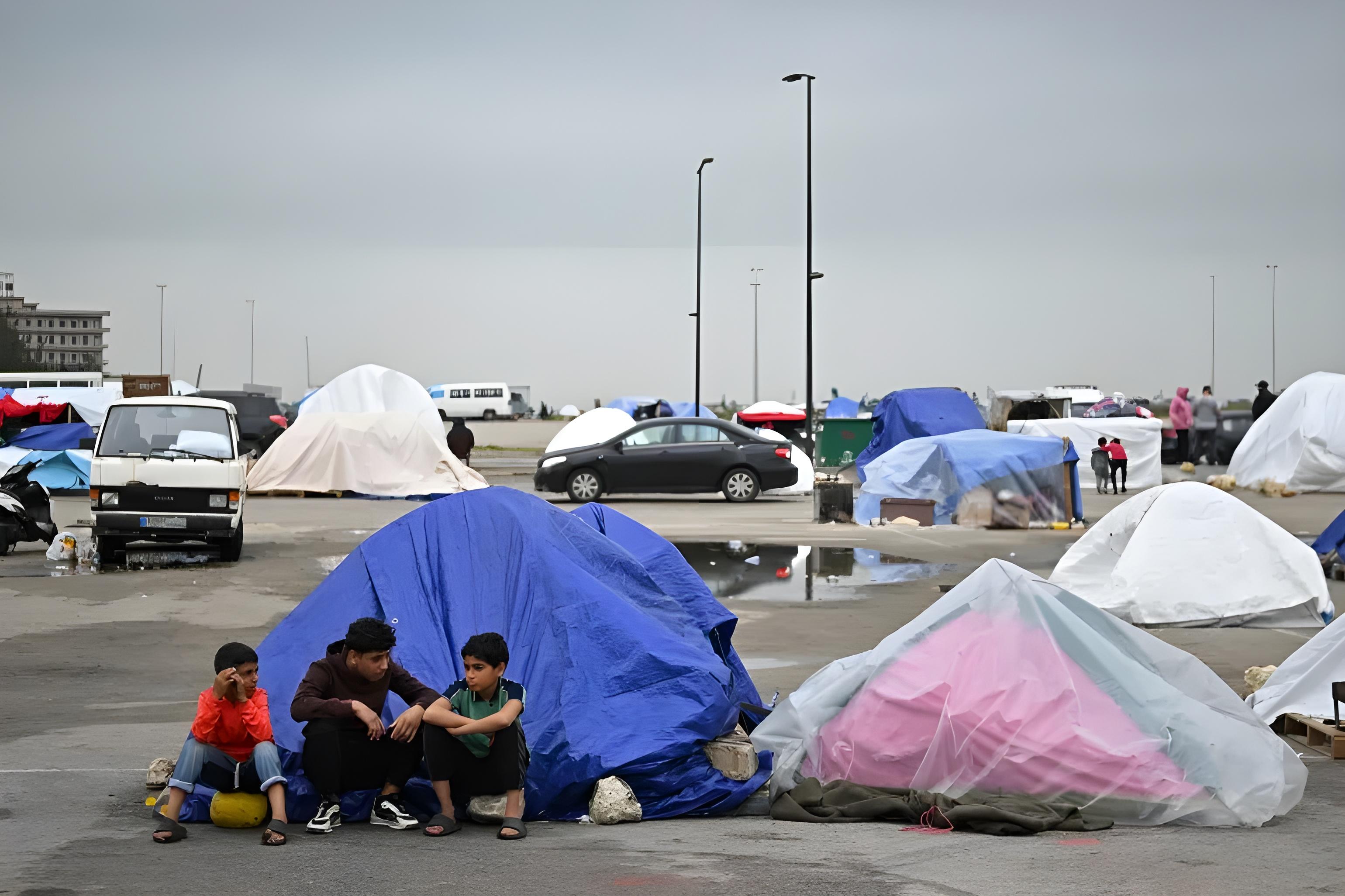 Trois enfants assis près d'une tente bleue dans un site de déplacement sous la pluie au Liban.